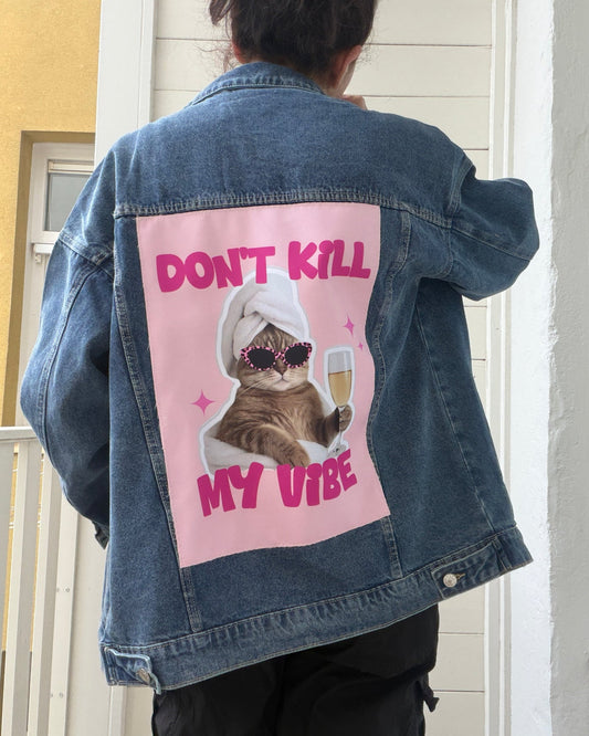 A person viewed from behind wearing an oversized blue denim jacket with a large pink graphic patch on the back. The patch features a cat wearing sunglasses and a white towel, holding a drink, with the text 'DON'T KILL MY VIBE' in pink.