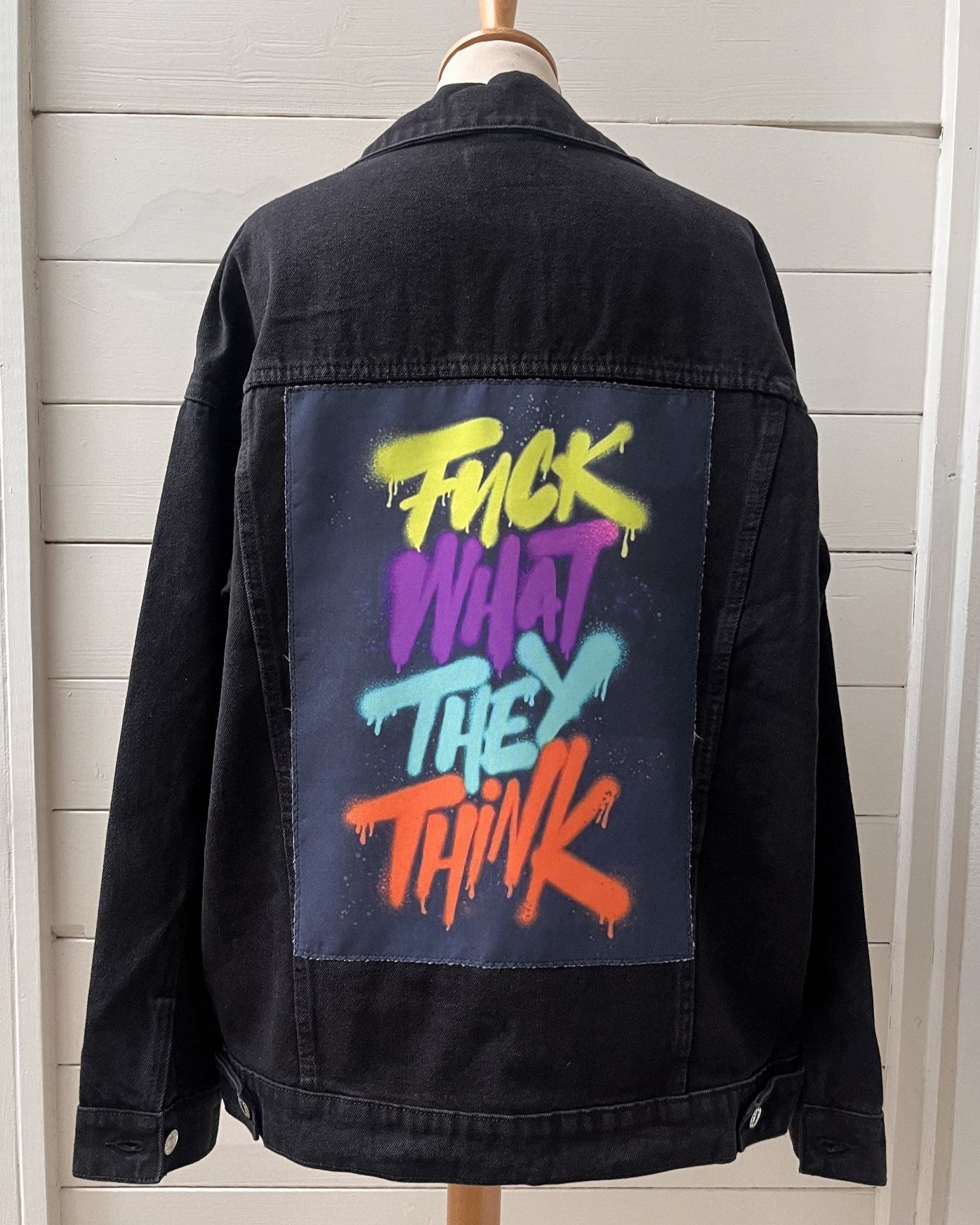 Black denim jacket with colorful text on the back, displayed on a mannequin against a light wooden panel background.