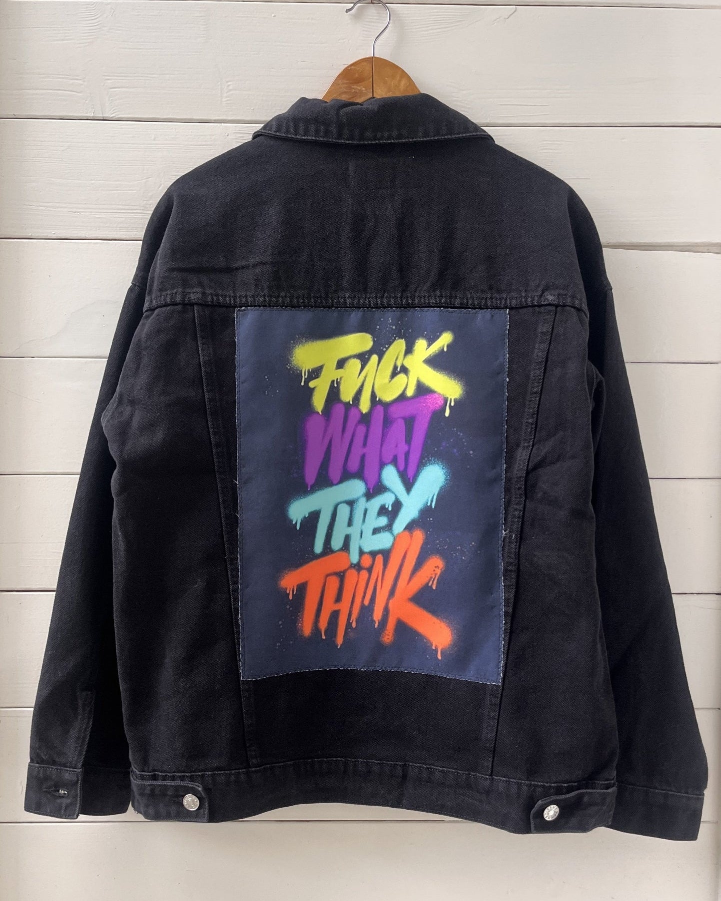 Black denim jacket with colorful text on the back, hanging against a wooden panel background.