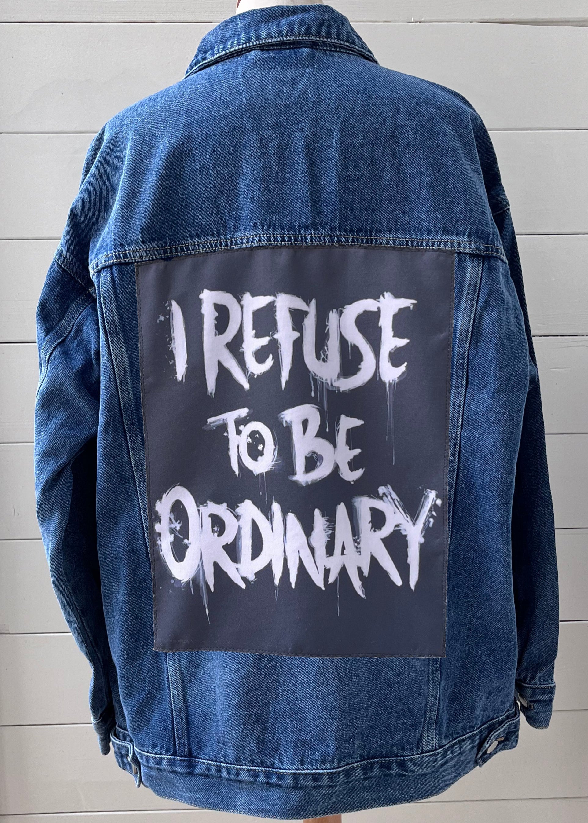 Blue denim jacket with 'I Refuse to be Ordinary' text on a wooden stand against a light gray background