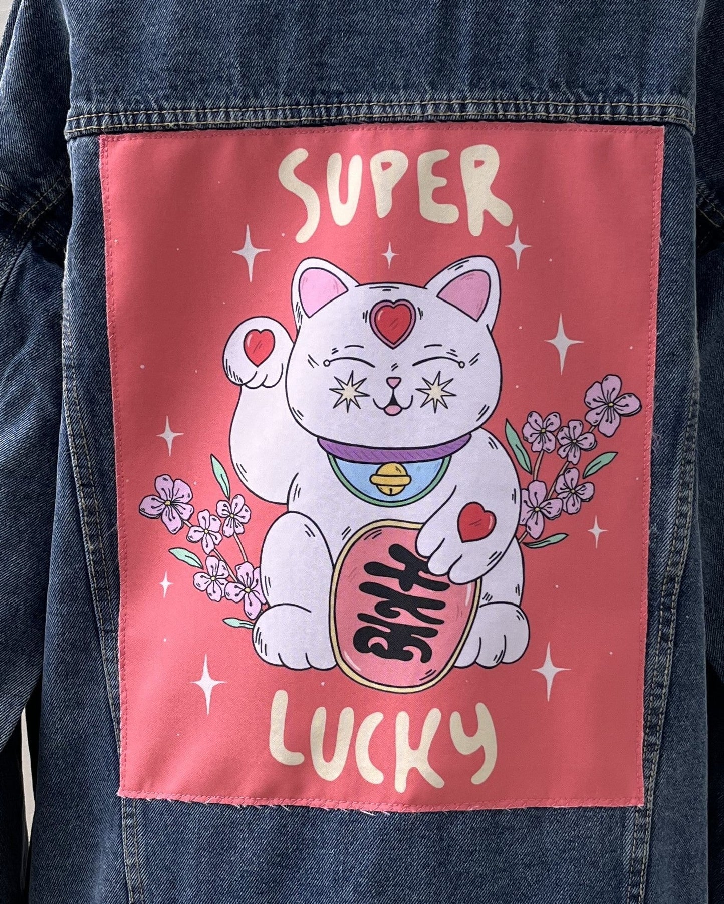 A back patch for a denim jacket featuring an illustration of a white cat with closed eyes, surrounded by floral elements and the text 'SUPER LUCKY'. The patch is black with a printed design.