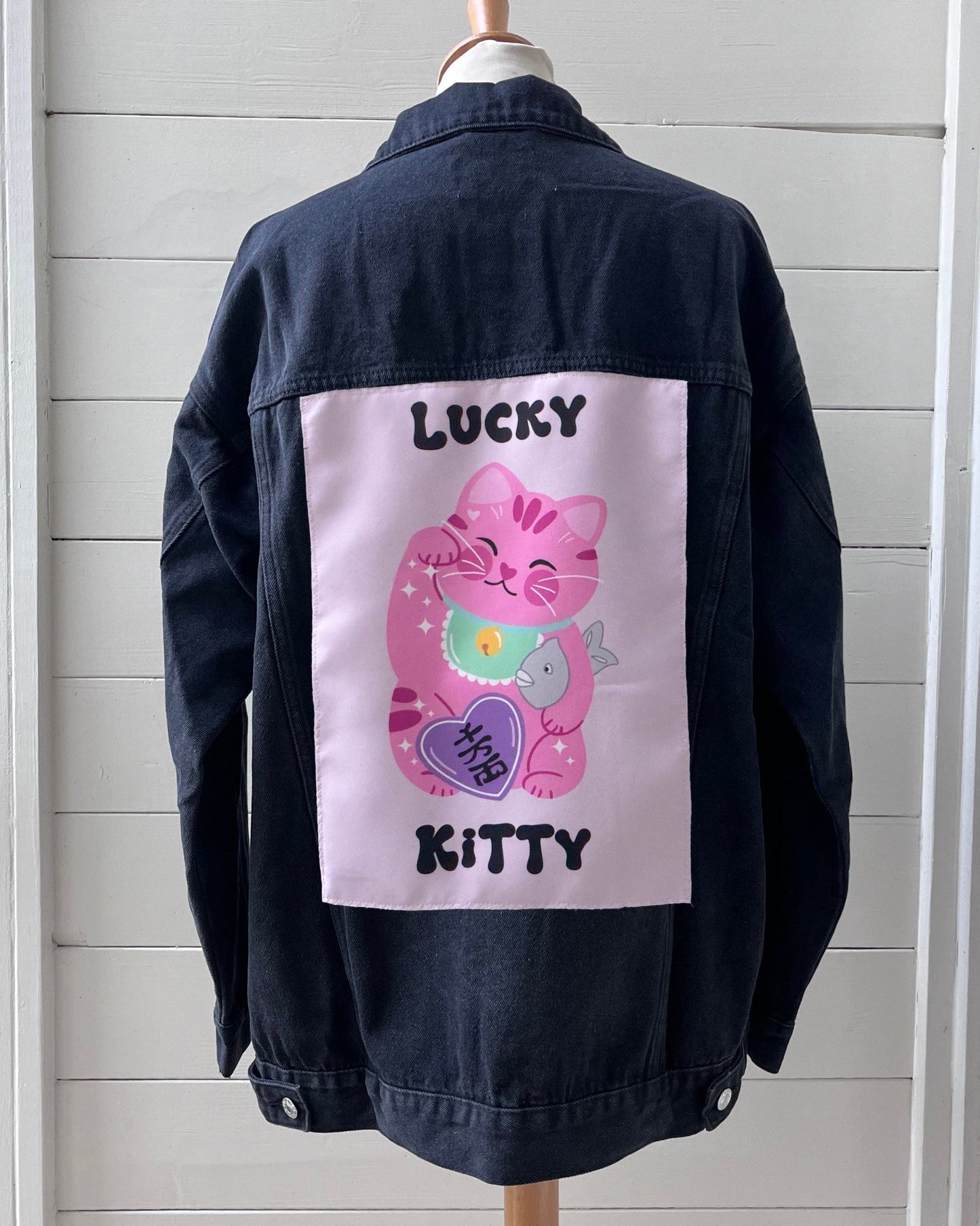 Black denim jacket with a pink cat graphic and text on a wooden mannequin against a light gray background