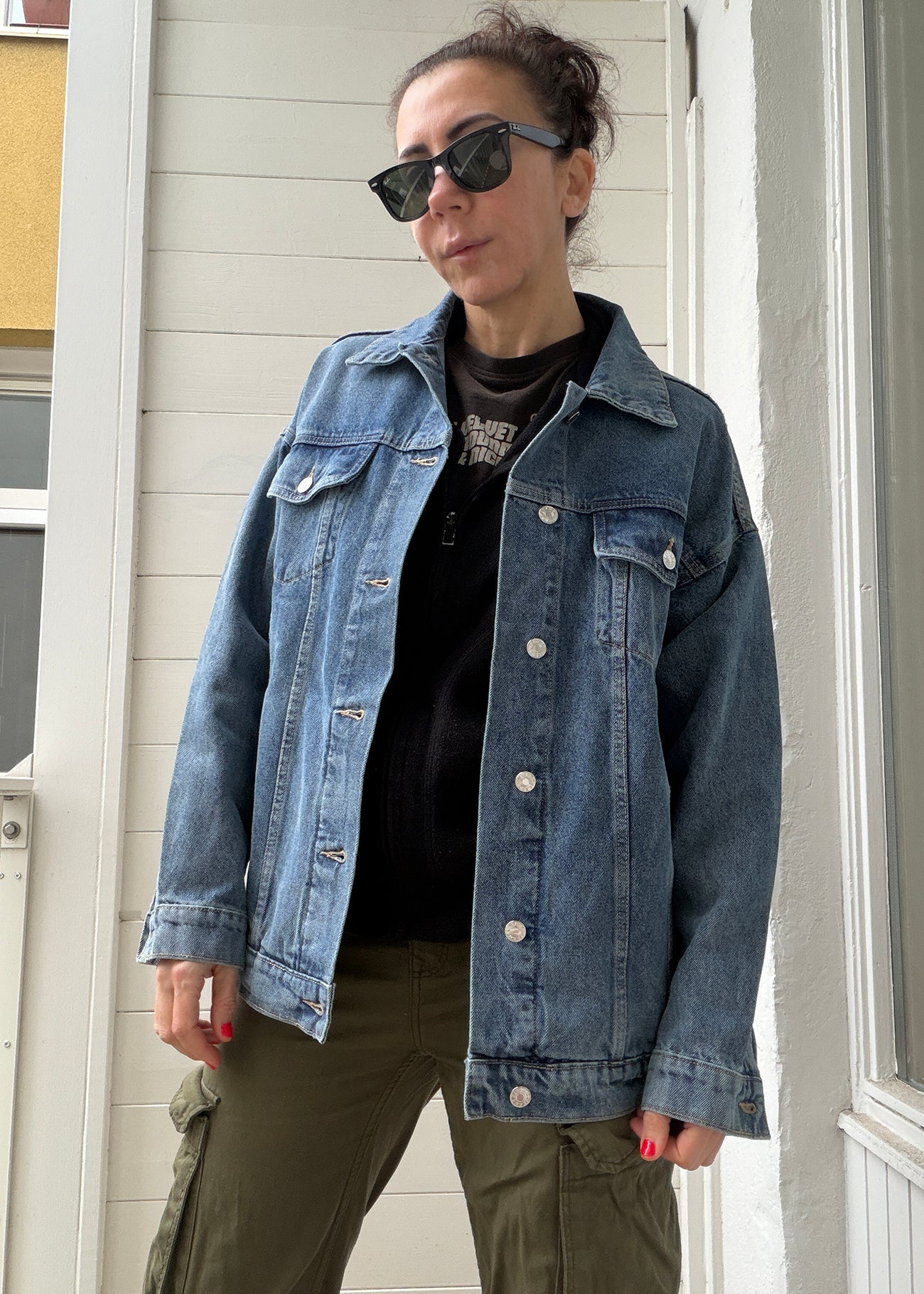 Person wearing a denim jacket and sunglasses standing in front of a white door.