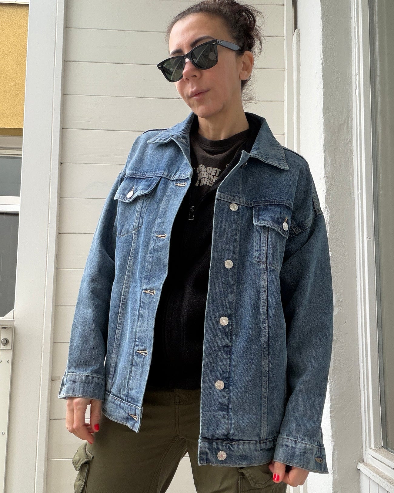Person wearing a denim jacket and sunglasses standing in front of a white door.