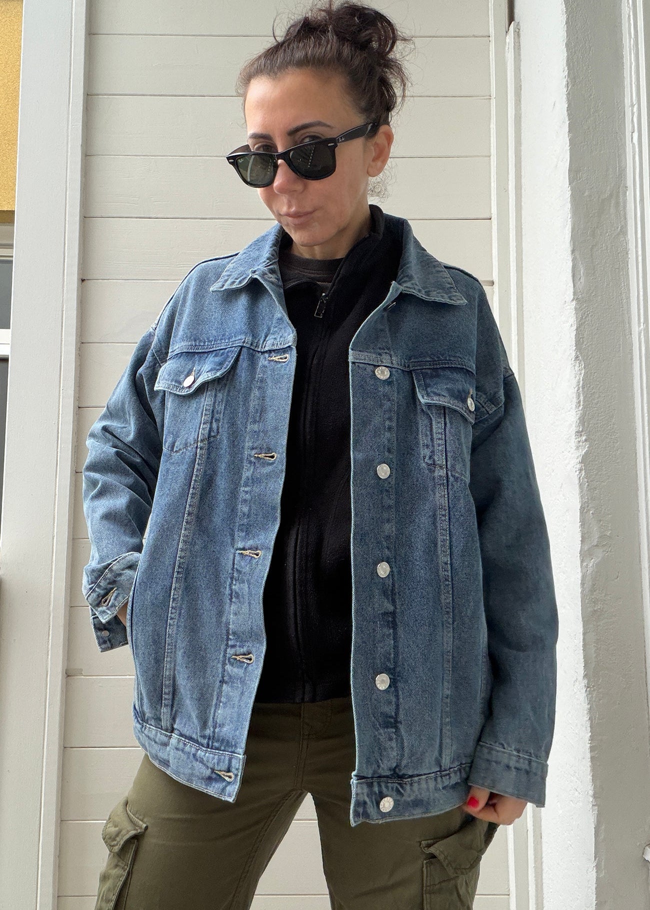 Person wearing a denim jacket and sunglasses standing against a white wall.
