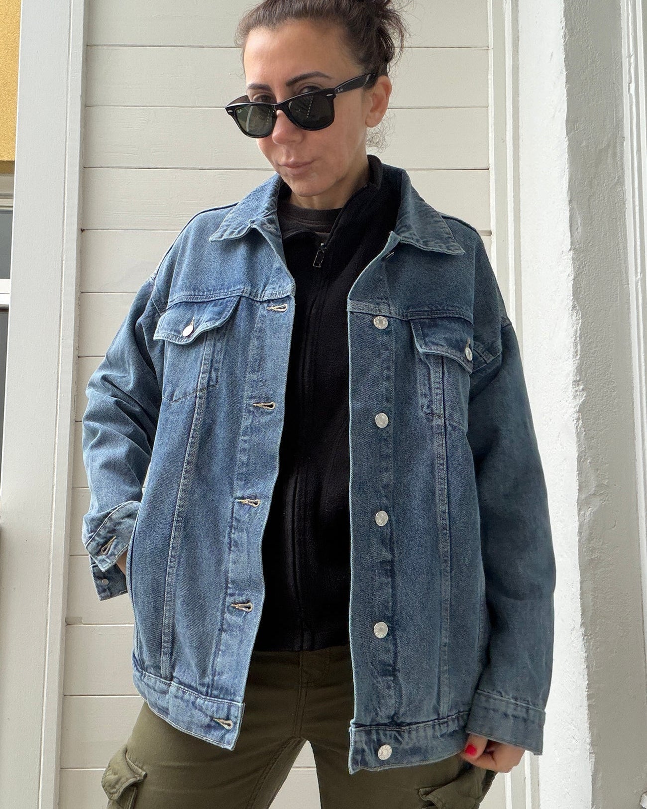 Person wearing a denim jacket and sunglasses standing against a white wall.