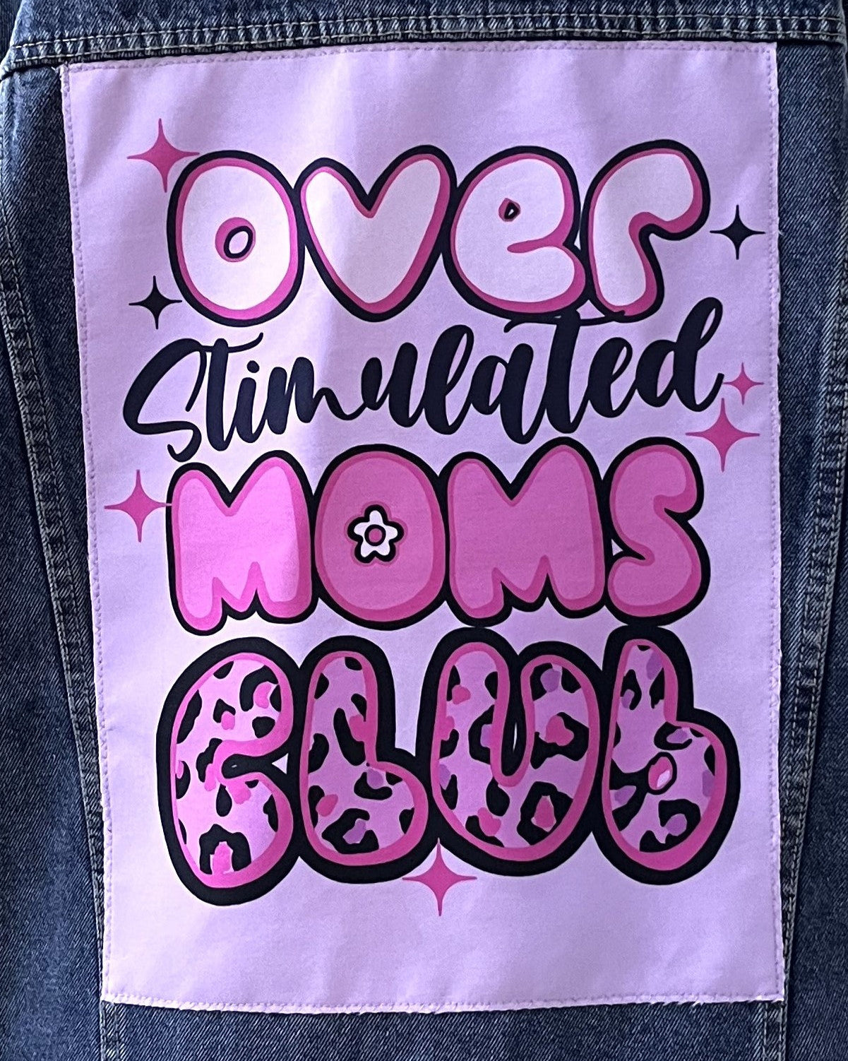Denim jacket with a patch reading 'Over Stimulated Moms Club' in pink and black text.