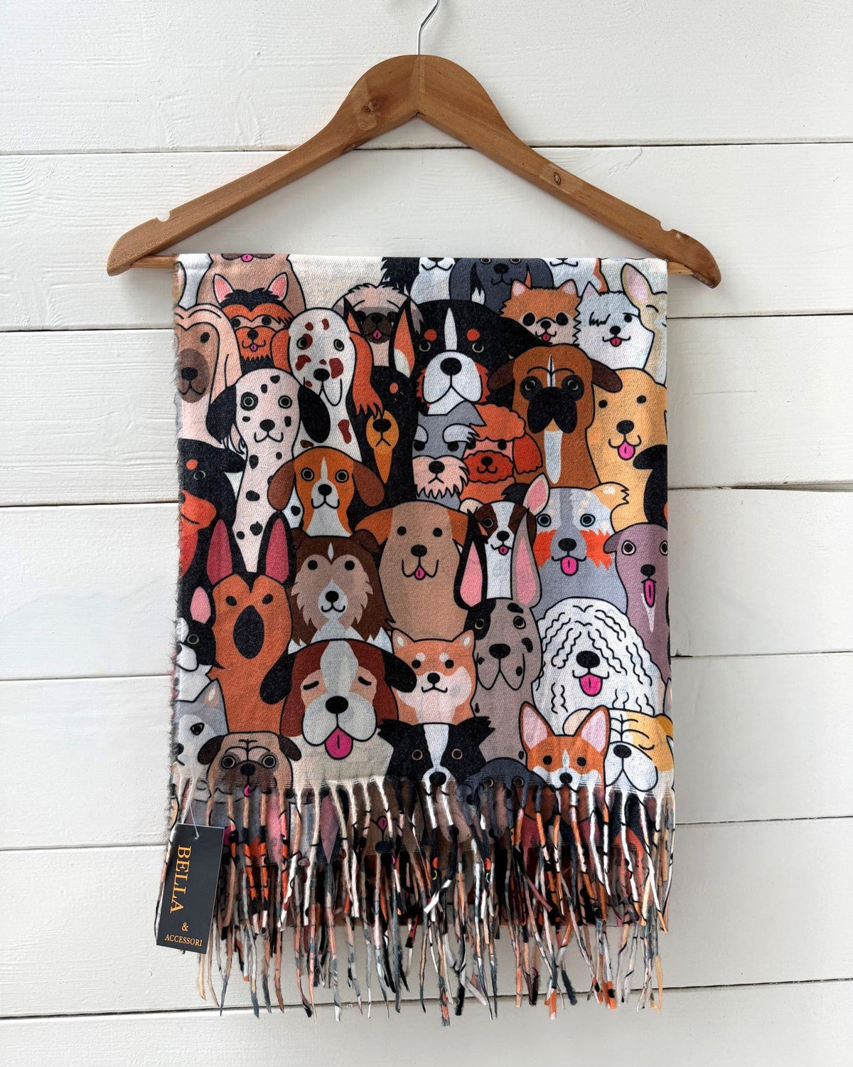 A fringed scarf featuring a dense cartoon illustration of various dog breeds, hanging from a wooden hanger against a white wood plank wall. A black fabric tag with gold lettering reading 'BELLA' is visible near the bottom left fringe.