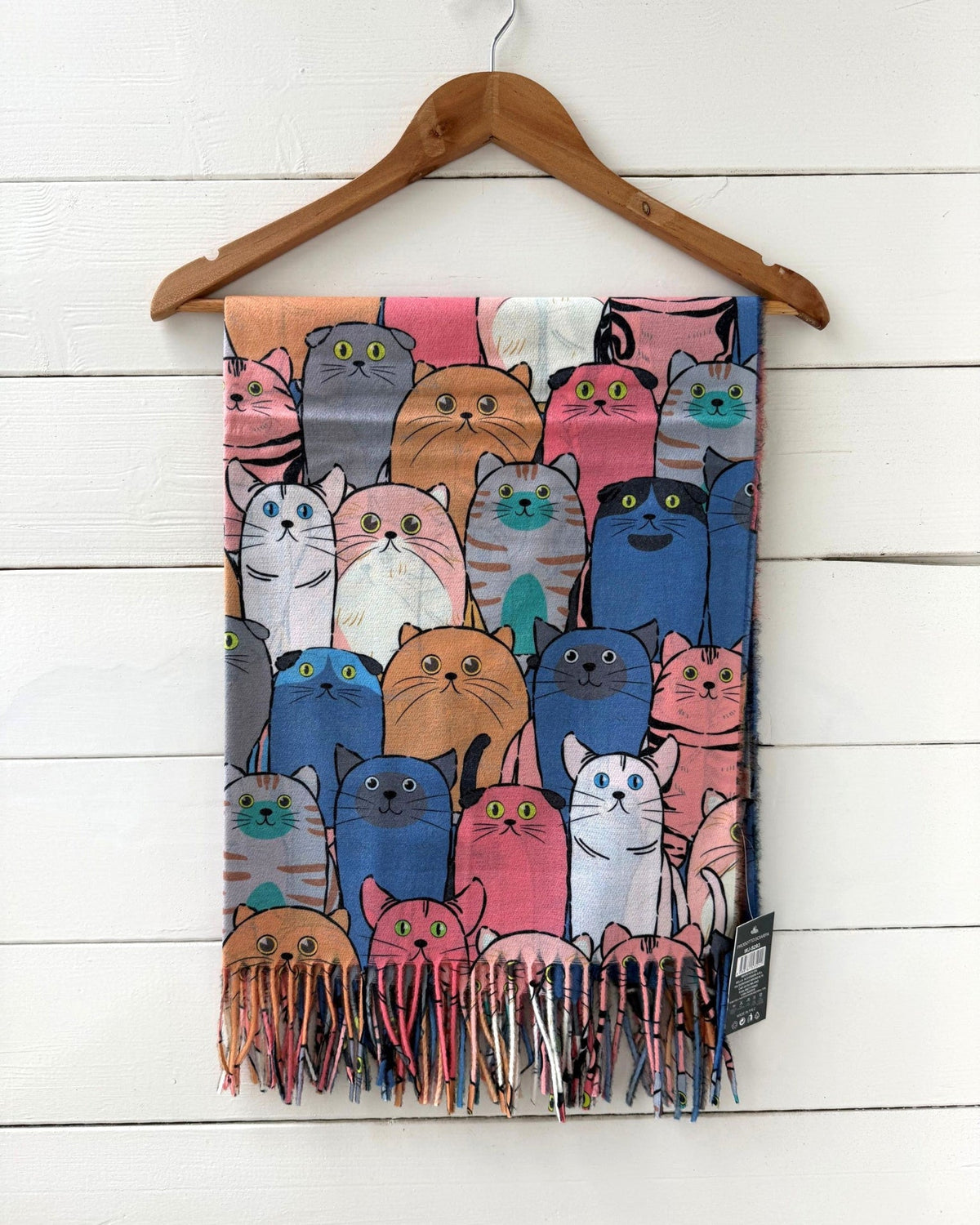 A fringed scarf featuring a colorful cartoon cat pattern, hanging from a wooden hanger against a white wood plank wall.