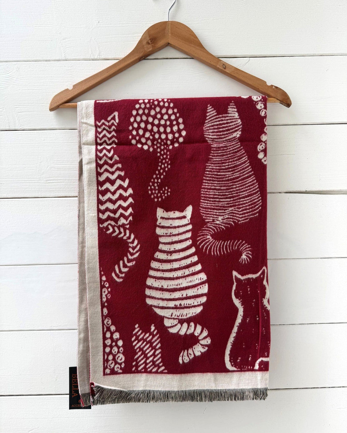 A rectangular scarf with fringe, featuring a deep red background and white cat illustrations, hanging from a wooden hanger against a white wood-paneled wall.