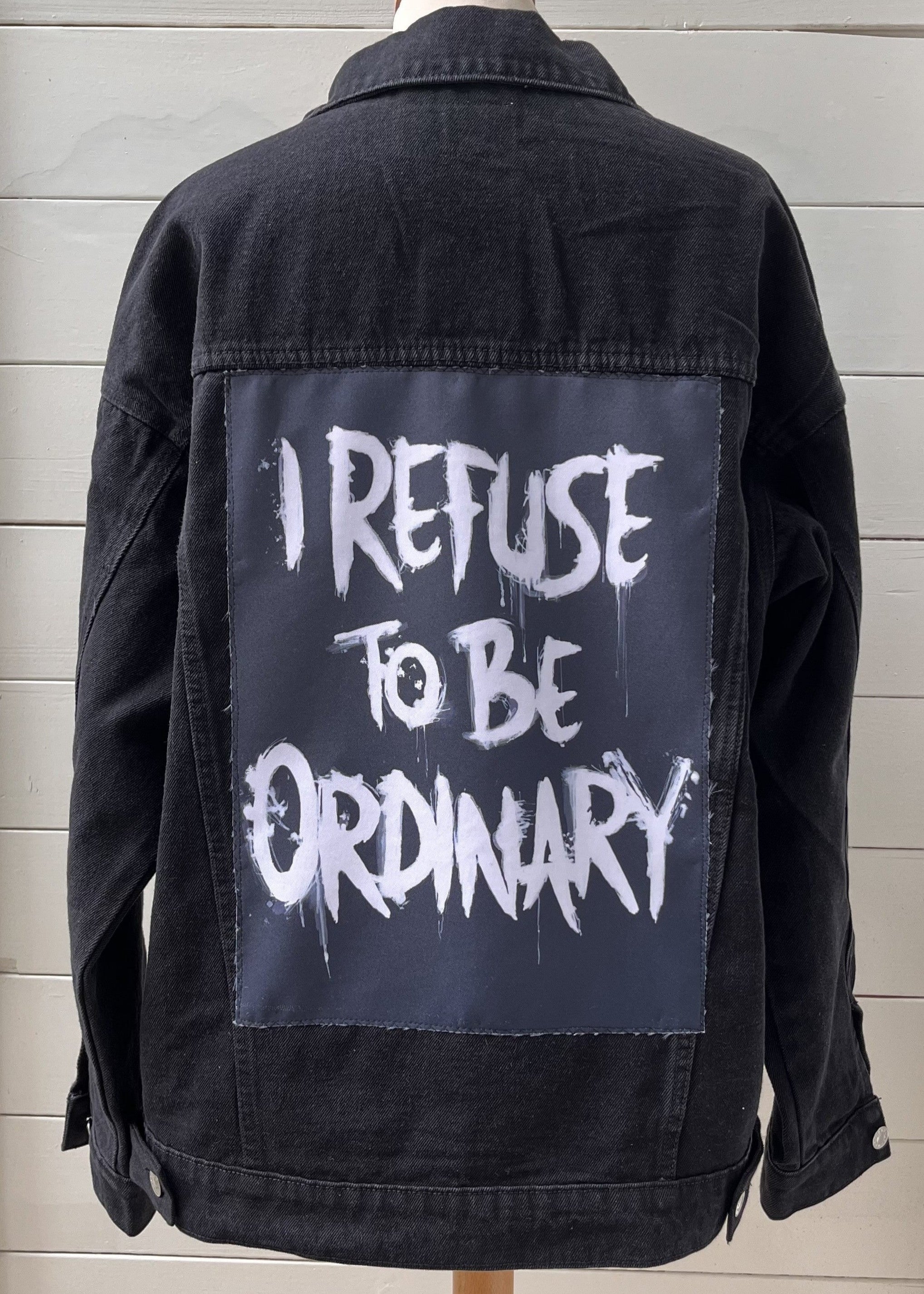Black denim jacket with 'I Refuse to be Ordinary' text on a wooden background