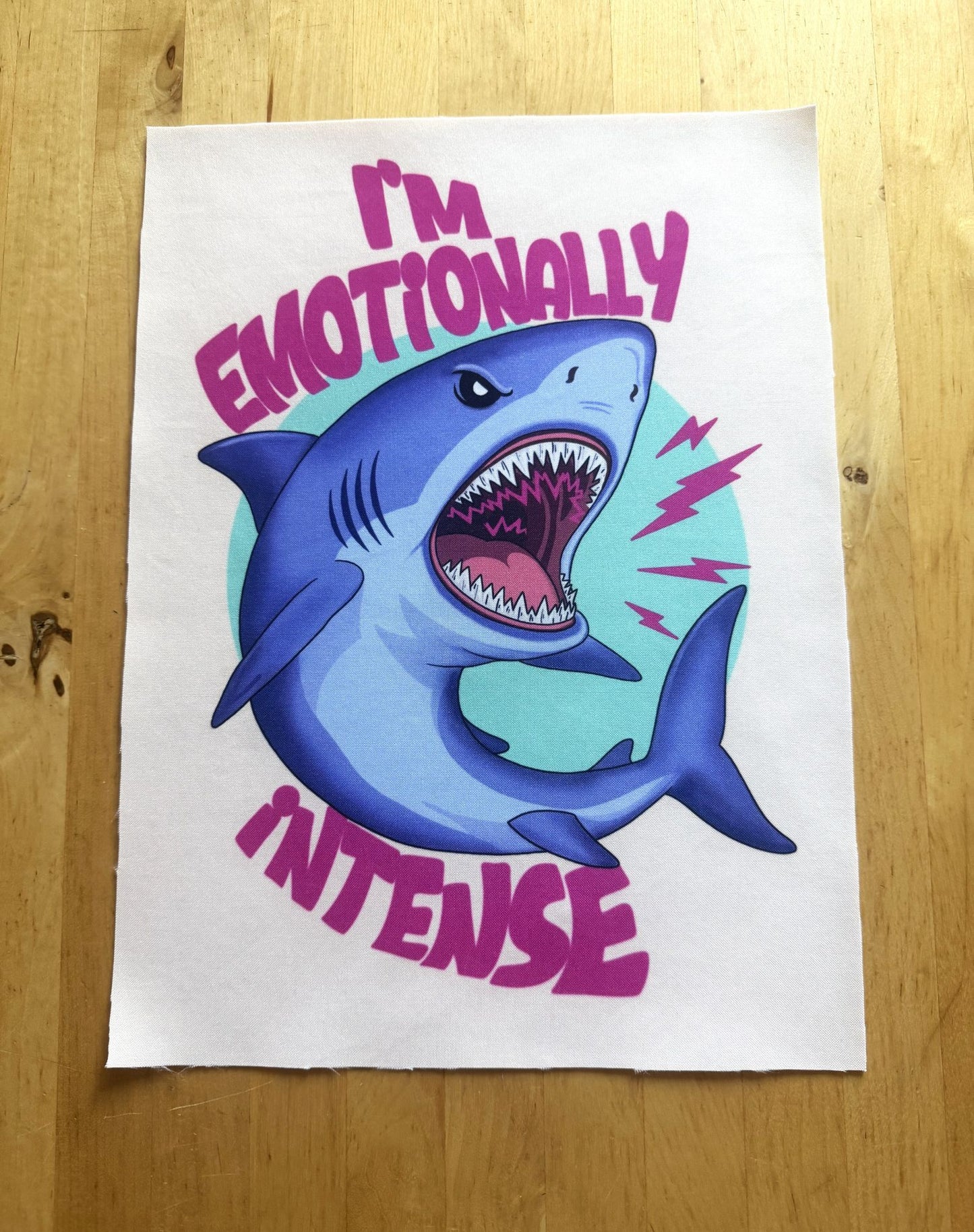 A white art print featuring a cartoon blue shark with its mouth wide open, surrounded by pink text that reads 'I'M EMOTIONALLY INTENSE' and a light teal circular background, placed on a wooden surface.