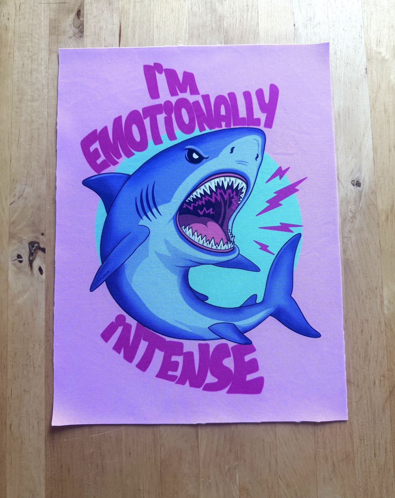A square art print featuring a cartoon shark with its mouth wide open, surrounded by the text 'I'M EMOTIONALLY INTENSE' in purple on a light purple background, displayed flat on a wooden surface.