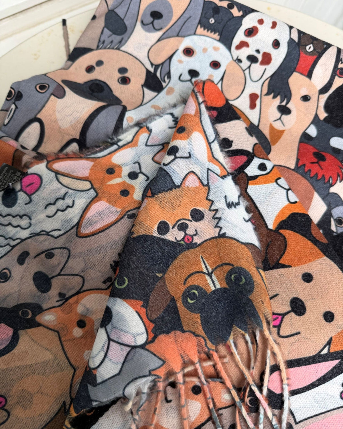 A close-up of a patterned scarf featuring cartoon dog faces in orange, brown, black, white, and gray, with fringed edges visible at the bottom.