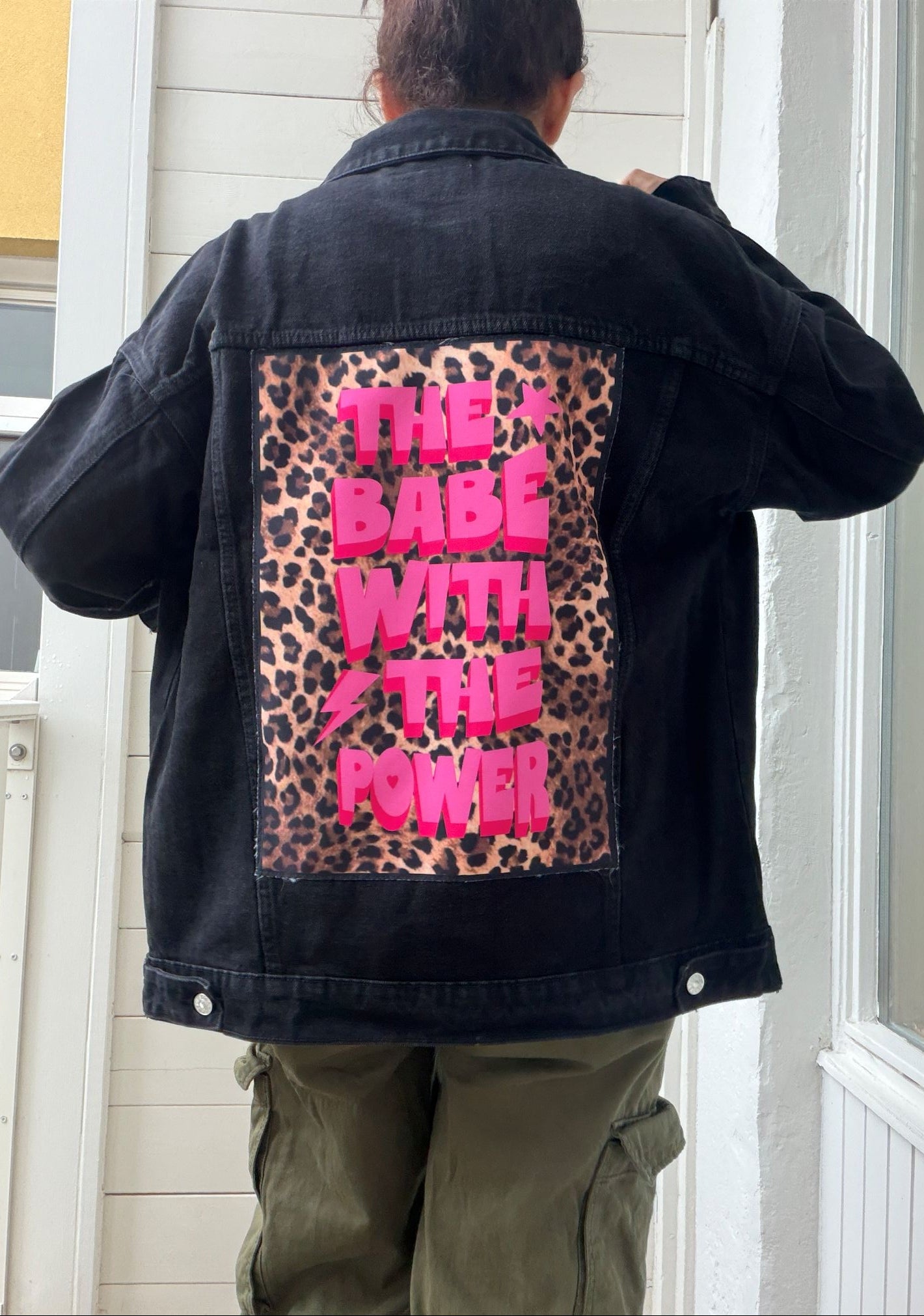 A person viewed from behind wearing a black denim jacket with a large back patch featuring a leopard print background and bright pink text that reads “THE BABE WITH THE POWER.”.