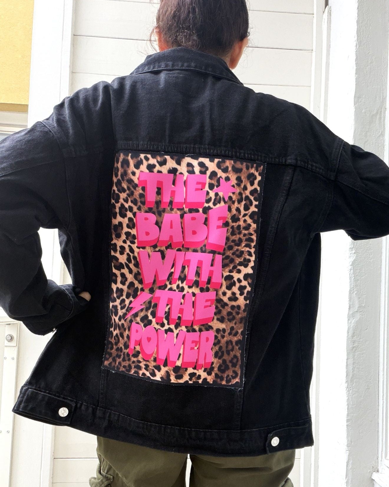 A person viewed from behind wearing a black denim jacket with a large back patch featuring a leopard print background and bright pink text that reads 'THE BABE WITH THE POWER'. The person is also wearing olive green cargo pants, standing in front of a white paneled door.