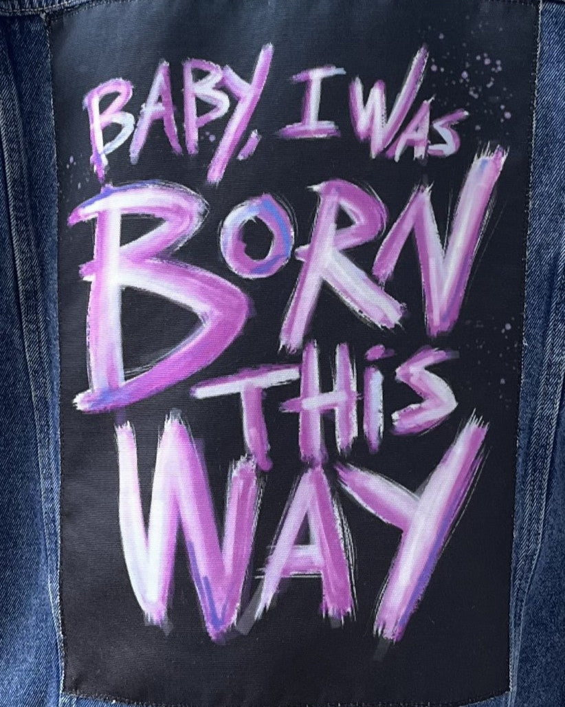 A black canvas patch with purple text 'BABY I WAS BORN THIS WAY' printed on it, designed for use on denim jackets or other fabrics.