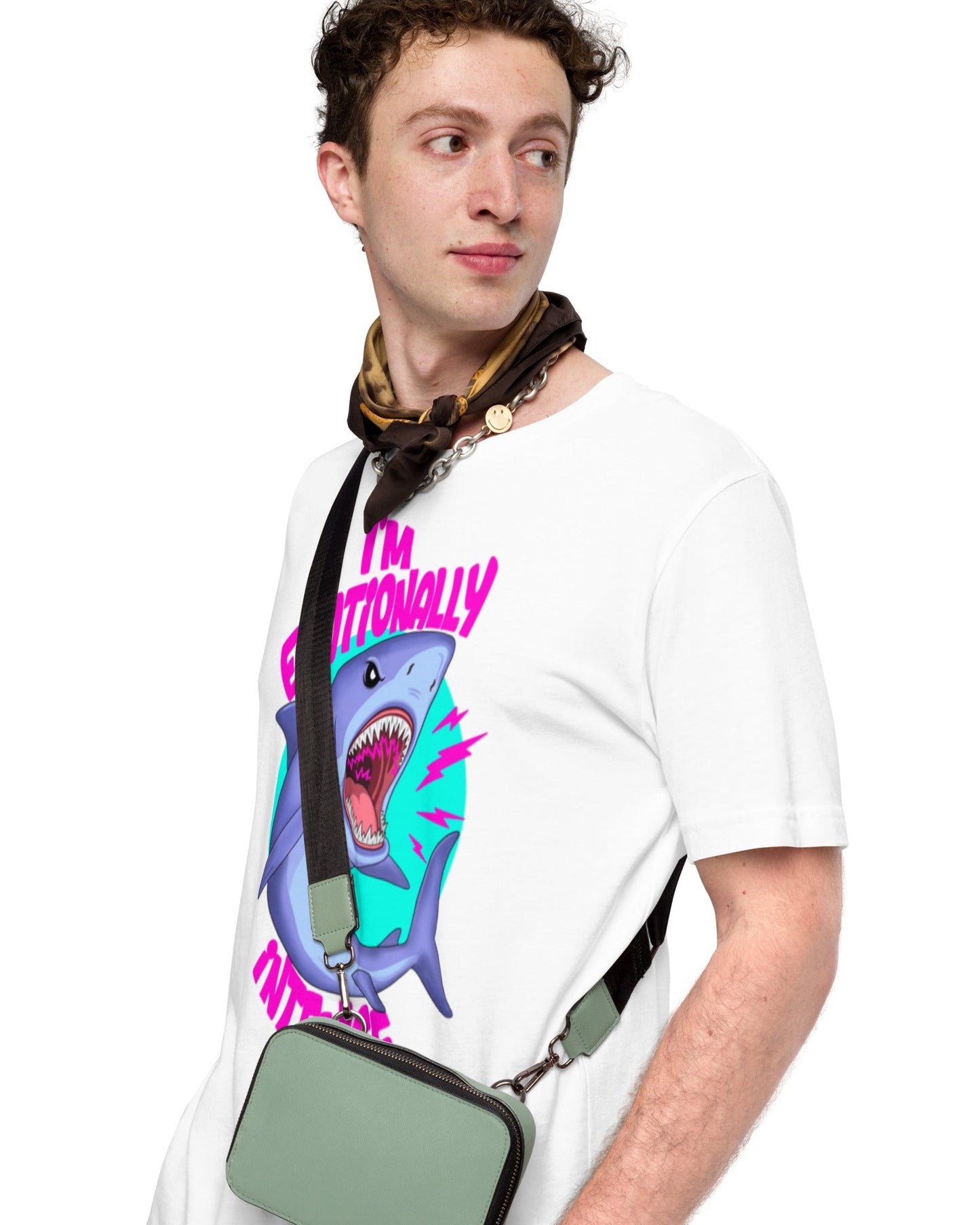 A man wearing a white graphic t-shirt with a colorful shark print and the text 'I'M EMOTIONALLY DEVASTATED'. He is also wearing a patterned neck scarf and a sage green crossbody bag with a black strap.