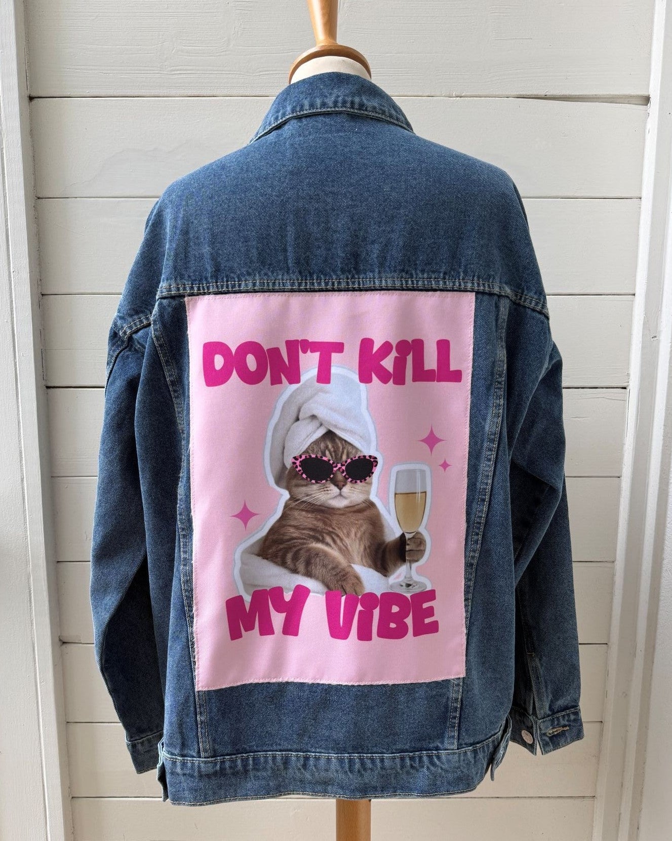 A blue denim jacket displayed on a wooden mannequin torso, showing the back with a large pink graphic patch featuring a cat in sunglasses and a towel with the text 'DON'T KILL MY VIBE'. The jacket is photographed against a white shiplap wall.