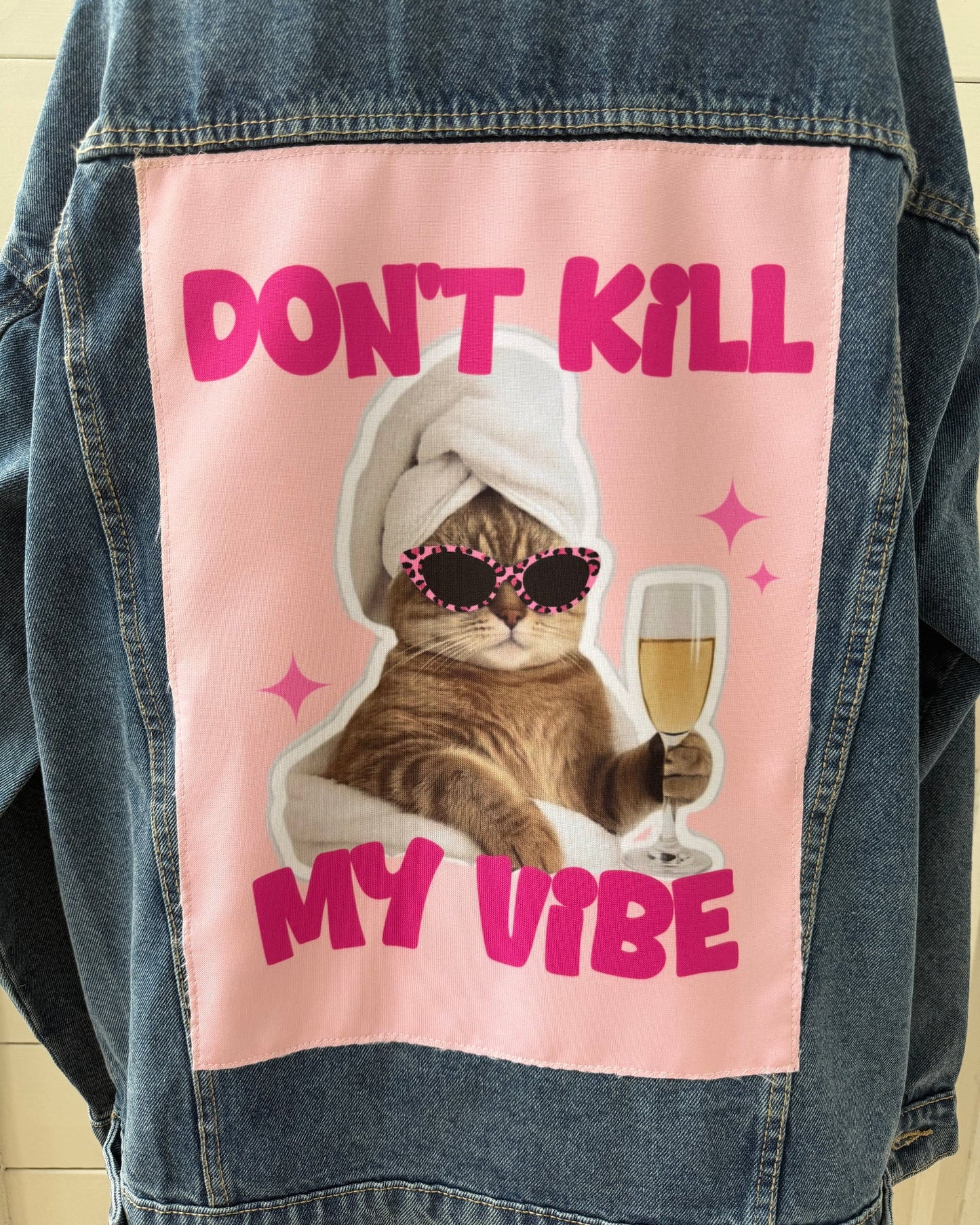 A denim jacket with a large pink graphic patch on the back featuring a cat in a white towel and sunglasses holding a champagne glass, with the text 'DON'T KILL MY VIBE'.