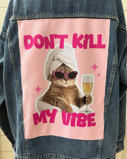 A denim jacket with a large pink graphic patch on the back featuring a cat in a white towel and sunglasses holding a champagne glass, with the text 'DON'T KILL MY VIBE'.